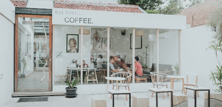 Outside of a white cafe with a sign saying "but first coffee".