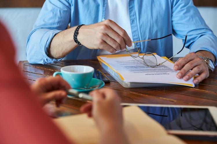 Two people sit across from each other with paperwork, and coffee.