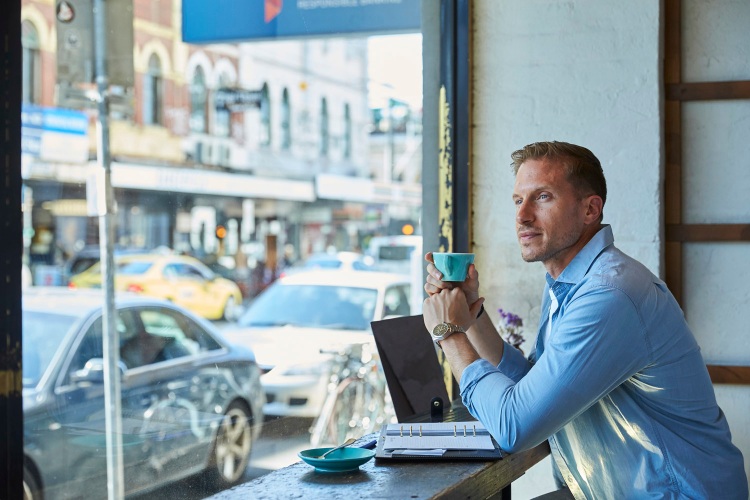 Man holding a coffee sits with his laptop in the window of a cafe overlooking a busy road.