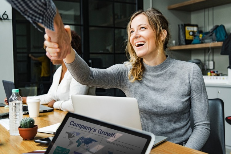 Smiling lady shaking hands with another person over a laptop screen.