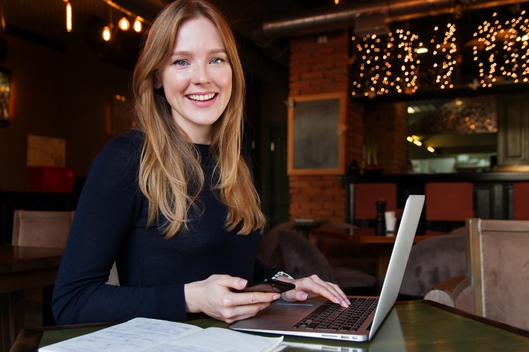 A women sits in a cafe with a computer and cell phone, looks directly at the camera.